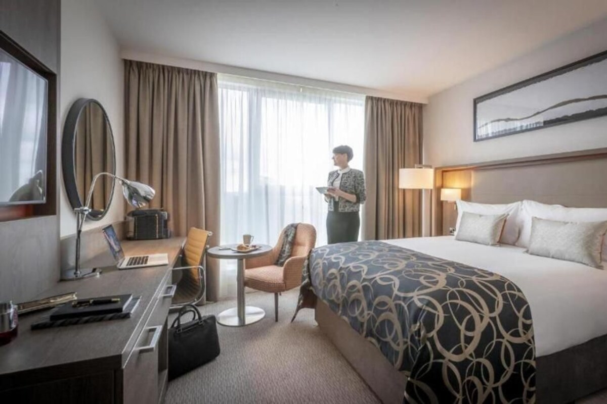 Ein Hotelzimmer mit einem großen Bett, einem Schreibtisch mit einem Laptop, einem Sessel, einer Lampe und einem Fenster mit Vorhängen.