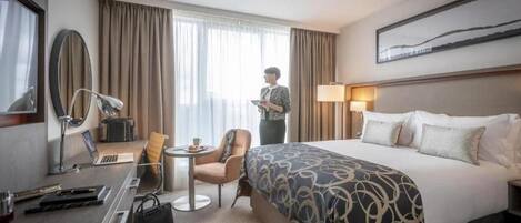 Una habitación de hotel con una cama grande, un escritorio con una computadora portátil, una silla, una lámpara y un ventanal con cortinas.