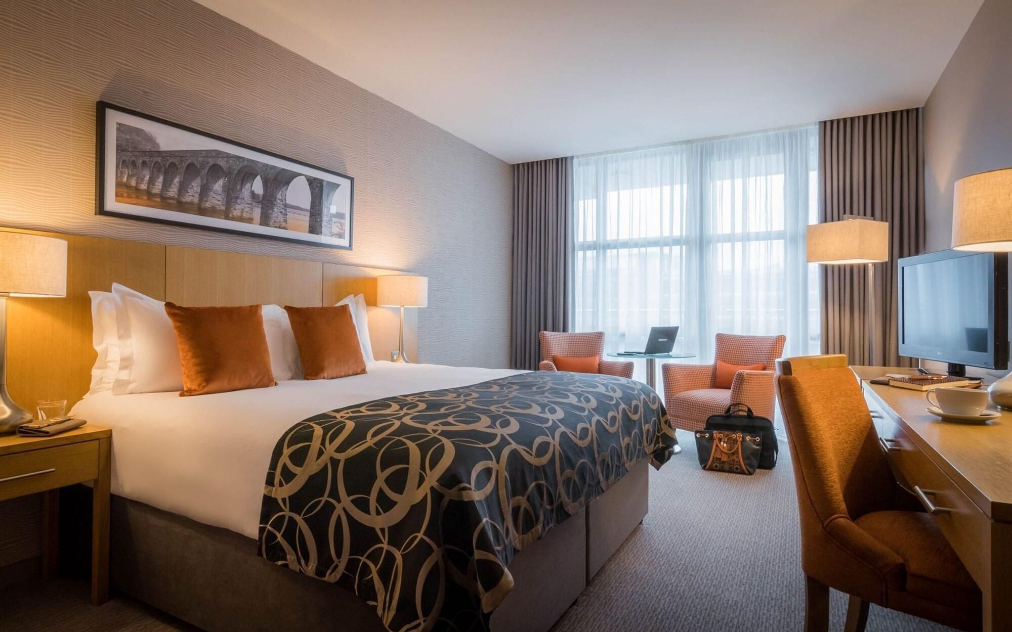 A hotel room with a large bed, two bedside tables, a desk with a chair, a television, and a window with curtains.