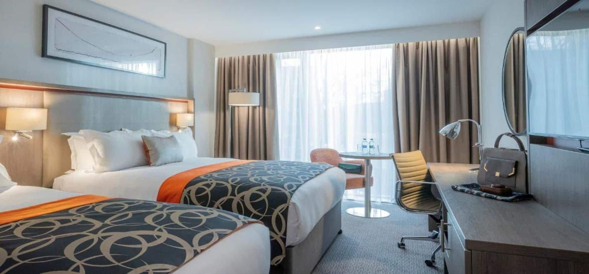 Une chambre d’hôtel avec deux lits, un bureau, une chaise, une lampe et une grande fenêtre avec des rideaux.