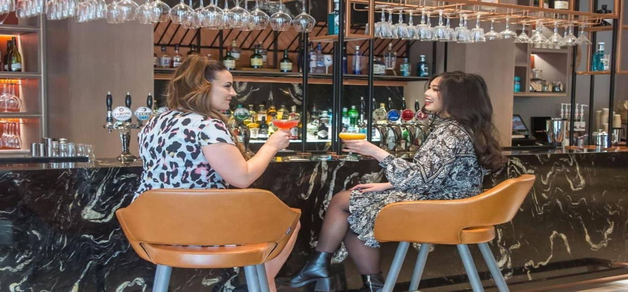 Two individuals seated at a bar with a variety of bottles and glasses in the background.