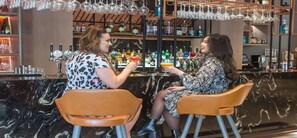Twee personen zitten aan een bar, met diverse flessen en glazen op de achtergrond.