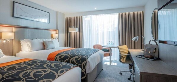 Habitación de hotel con dos camas, un escritorio, una silla, una lámpara y un ventanal grande con cortinas.