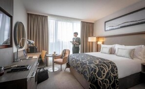 Une chambre d’hôtel avec un grand lit, un bureau avec un ordinateur portable, une chaise, une lampe et une fenêtre avec des rideaux.