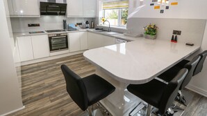 A modern kitchen with white cabinets, a central island with bar stools, stainless steel appliances, and a window with a view. - Bwthyn Eirlys (Cemaes Bay)