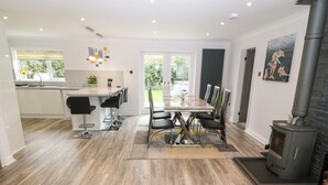 A modern kitchen with a central island, dining area with chairs, a large window, and a wood-burning stove. - Bwthyn Eirlys (Cemaes Bay)
