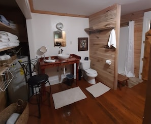 Une petite salle de bain avec un meuble-lavabo en bois, des toilettes et un lavabo. Des serviettes sont accrochées au mur, il y a un petit miroir et un tableau encadré au-dessus du miroir.