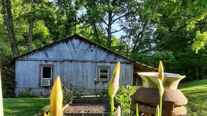 A rustic wooden barn with a metal roof, surrounded by lush greenery and flowering plants. - Whispering Waters Cabin by The Creek (Fairview)