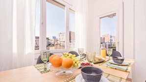 Taula de petit esmorzar amb fruita, un got de suc d'aranja i una ampolla d'aigua, situada en una habitació amb finestres grans que donen a un paisatge urbà.