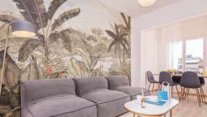 Un saló amb un sofà seccional gris, una taula de centre rodona i un mural amb vegetació tropical a la paret.