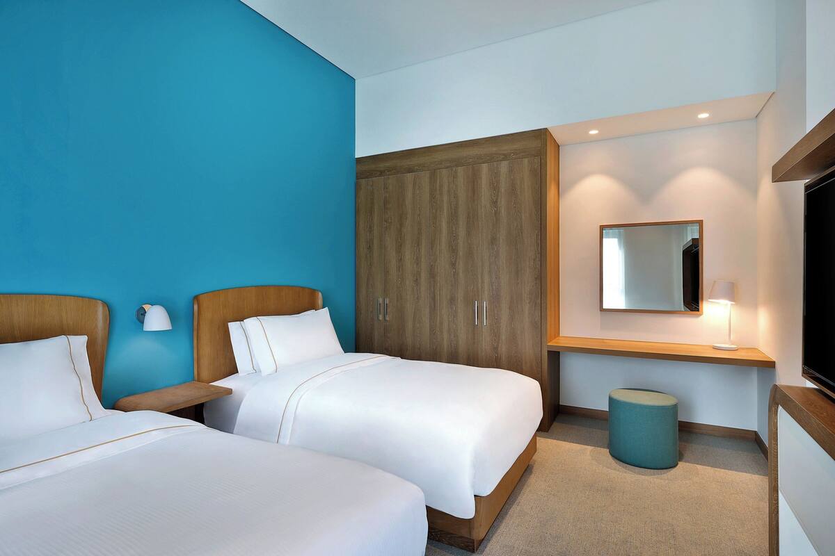 A hotel room with two beds, a wooden wardrobe, a mirror, a lamp, and a television.