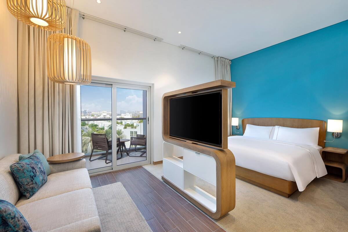 A hotel room with a large bed, a TV, a sofa, and a balcony with a view of the city.