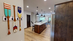 A modern kitchen with wooden cabinets, a central island, pendant lighting, and a view to a dining area. - The Blue Ox on Gull, 5 Bed 5 Bath Resort Home, Sleeps 17 with Boat Slip on Gull Lake (Lake Shore)