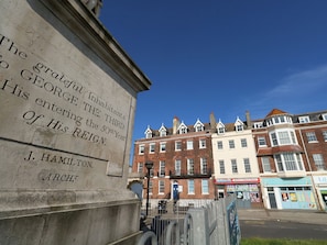A stone monument with inscriptions, a street view with buildings, and a clear blue sky. - The Kings Statue (Weymouth)