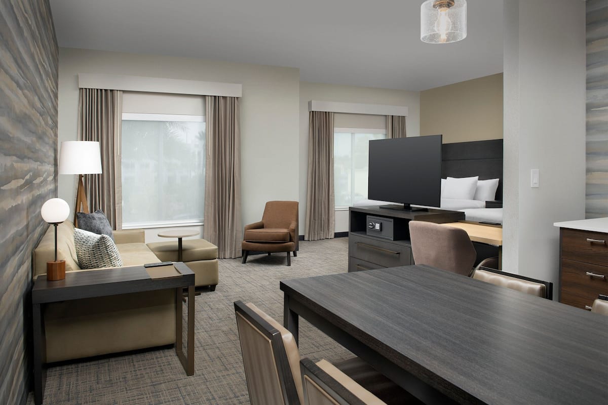 Una habitación de hotel moderna con sofá, sillón, mesa de comedor, televisión y escritorio con lámpara.