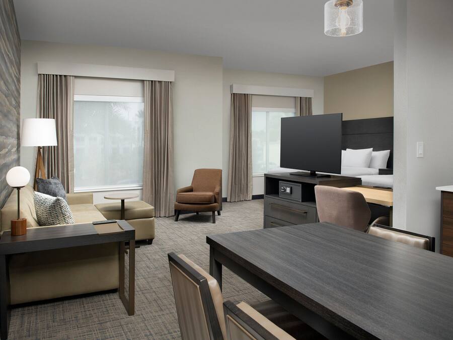 A modern hotel room with a sofa, armchair, dining table, TV, and a desk with a lamp.