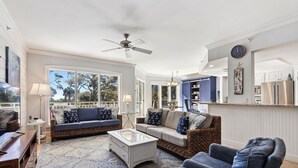 Ein geräumiges Wohnzimmer mit großem Fenster, Deckenventilator und einer Küchenzeile mit blauen Schränken.