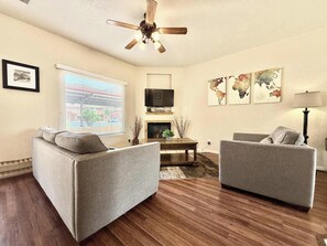 Un salon avec deux canapés gris, un plancher en bois, un ventilateur de plafond, un téléviseur et une cheminée.