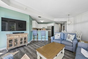 Un salon avec un canapé, une table basse et un téléviseur, une cuisine avec des tabourets de bar et un coin repas avec des chaises.