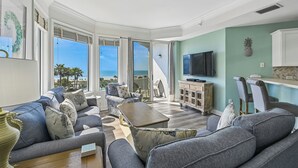 Un salon spacieux avec un grand canapé, une table basse et un téléviseur. Il y a un balcon avec vue sur l’océan.