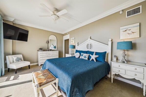 A bedroom with a bed, a chair, a dresser, a nightstand, a ceiling fan, and artwork on the walls.