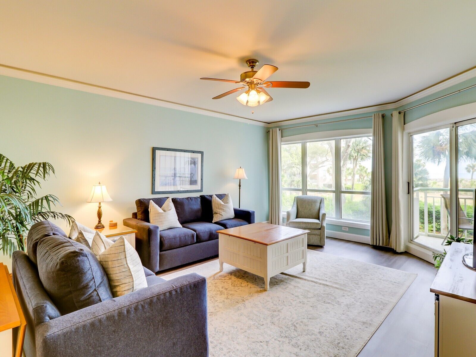 A spacious living room with a grey sofa, a white coffee table, and a ceiling fan. There is a window with a view of palm trees.
