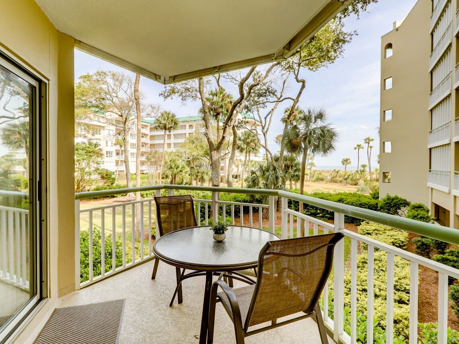 A balcony with a table and chairs, overlooking a landscaped area with trees and a distant building.