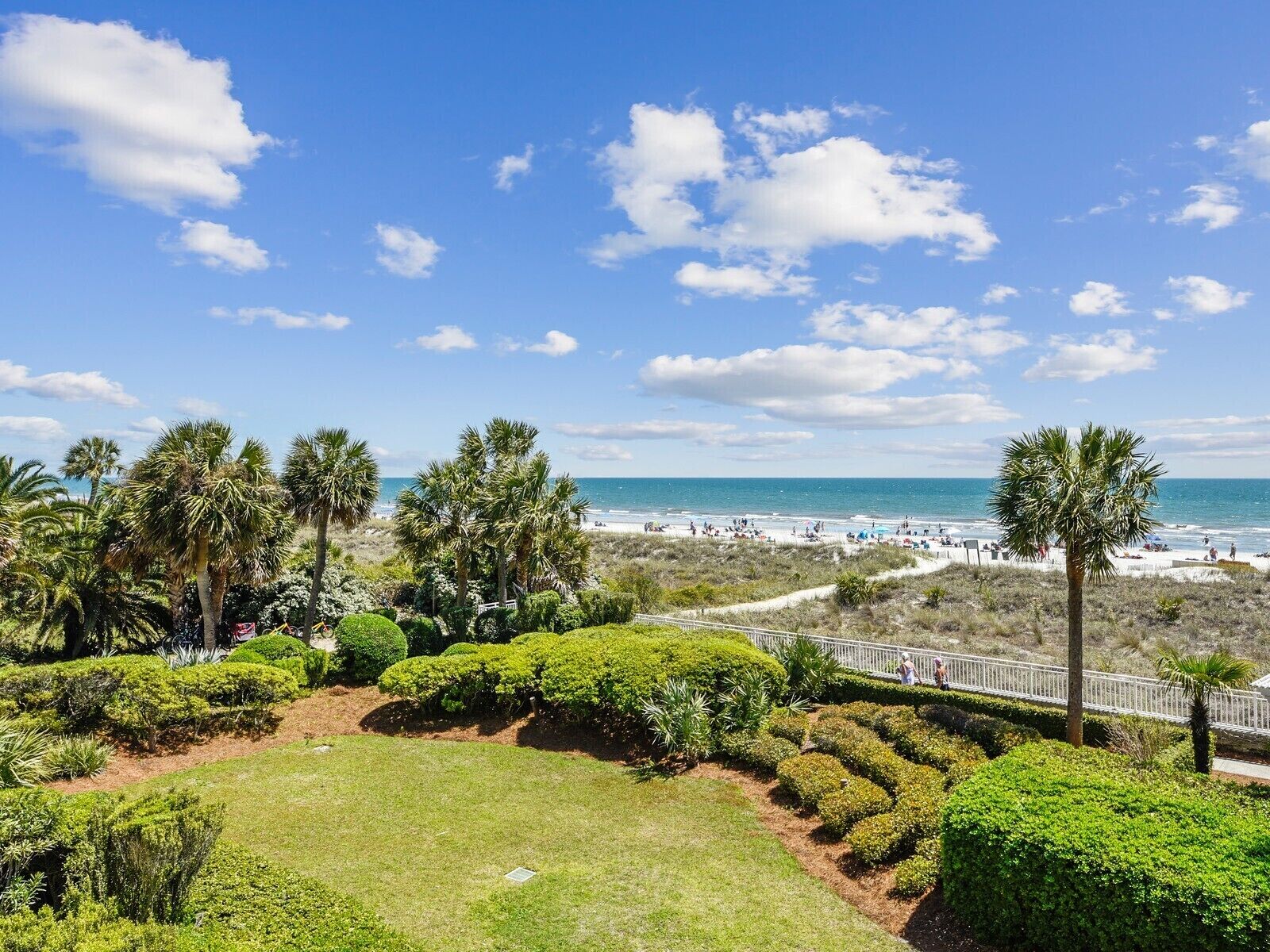 A landscaped garden with manicured bushes and palm trees, a pathway, and a view of the beach and ocean.