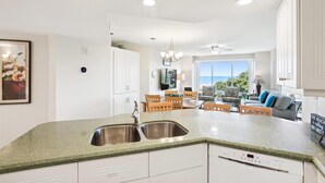 A modern kitchen with a granite countertop, stainless steel sink, and white cabinetry. There is a dining area with wooden chairs and a sofa, and a large window offering a view of the ocean.
