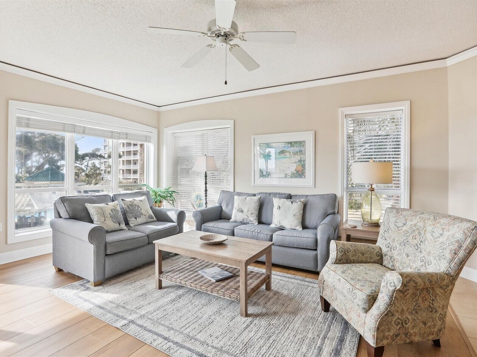 A spacious living room with two grey sofas, a patterned armchair, a wooden coffee table, and a ceiling fan. Large windows provide natural light and views of the outside.