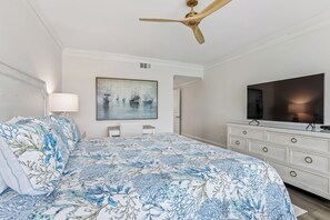 A bedroom with a bed, a ceiling fan, a painting, a dresser, and a television.