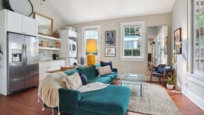 A modern living room with a teal sofa, a glass coffee table, and a kitchen area with a refrigerator and washing machine.
