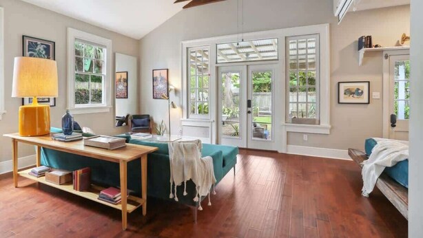 A spacious room with a wooden floor, a teal sofa, a wooden table with books, a lamp, and a window with a view of greenery.
