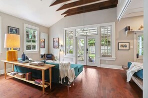 Een ruime kamer met een houten vloer, een blauwgroene bank, een houten tafel met boeken, een lamp en een raam met uitzicht op groen.
