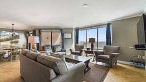 Ein geräumiges Wohnzimmer mit einer großen Couch, Sesseln, einem Couchtisch und einem Essbereich. Ein großes Fenster bietet Blick auf den Strand.