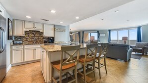Eine moderne Küche mit einer zentralen Kochinsel, ein Essbereich mit Stühlen und ein Wohnzimmer mit Sofa und Blick zum Himmel.