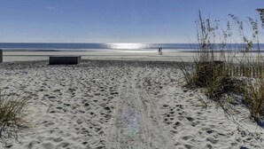 Une plage de sable avec des empreintes menant à l’eau, un ciel dégagé et une personne qui marche au loin.