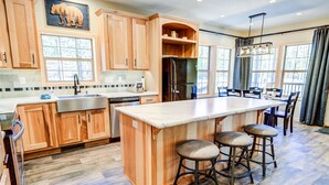 A spacious kitchen with wooden cabinets, a large island with stools, a refrigerator, and a dining area with chairs. - Shepherd Family Retreats- Bear Cub: 2BR, 2BA home 6 min from Yellowstone National Park (West Yellowstone)