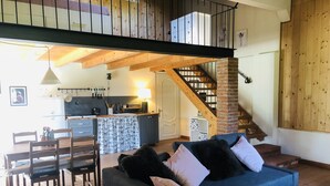 A cozy living room with a sofa, a dining table with chairs, a kitchen area with a staircase, and a wooden ceiling. - The house in the fields_Dépendance (Lombardia)