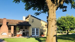 A house with a brick chimney, a tiled roof, a white door, and a large tree in front. - The house in the fields_Dépendance (Lombardia)