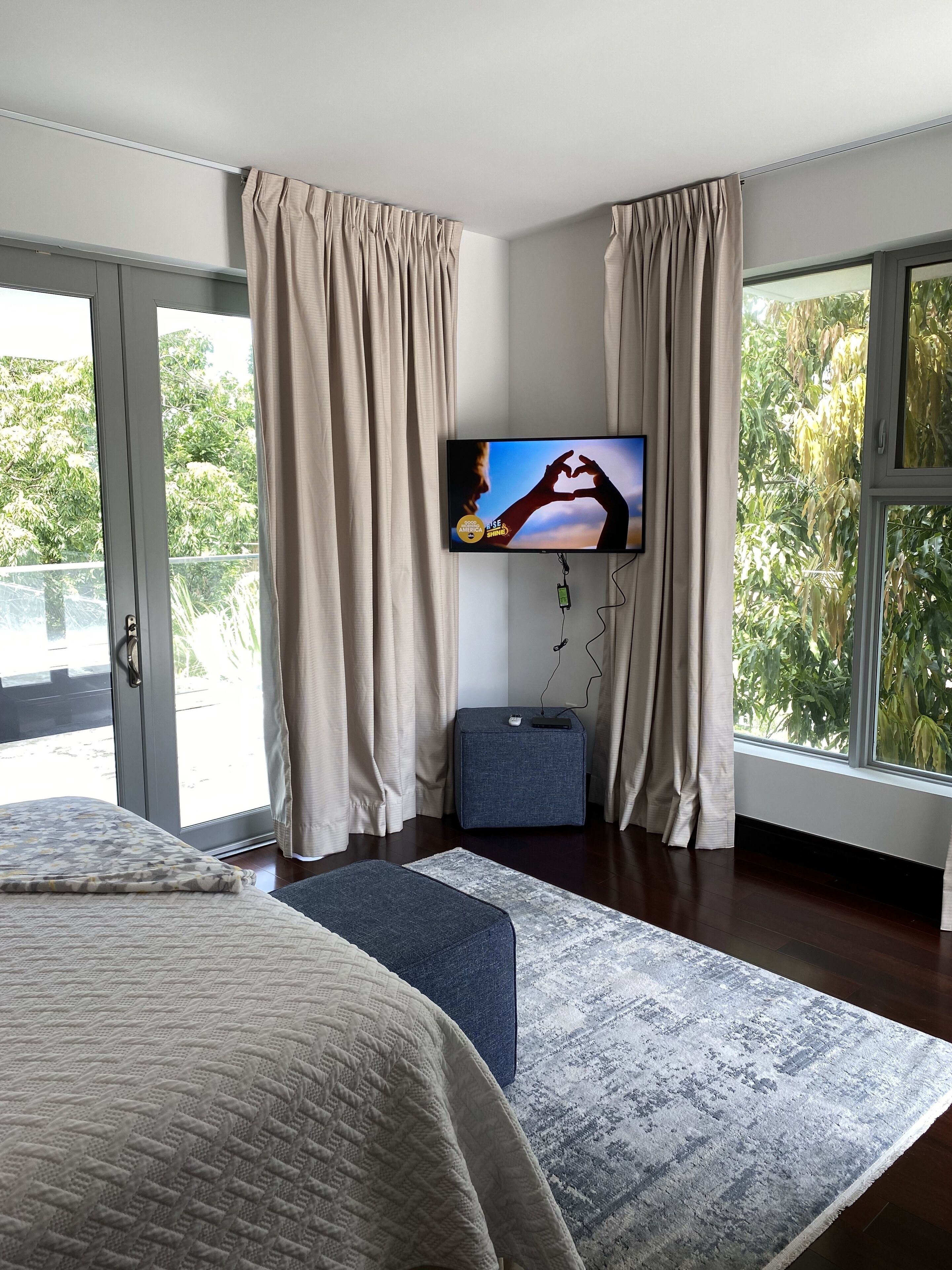 A modern bedroom with a large bed, a flat-screen TV mounted on the wall, a small side table, and a view of greenery through the windows.