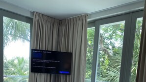 A hotel room with a large bed, a television mounted on the wall, a small seating area, and a balcony with a view of palm trees.