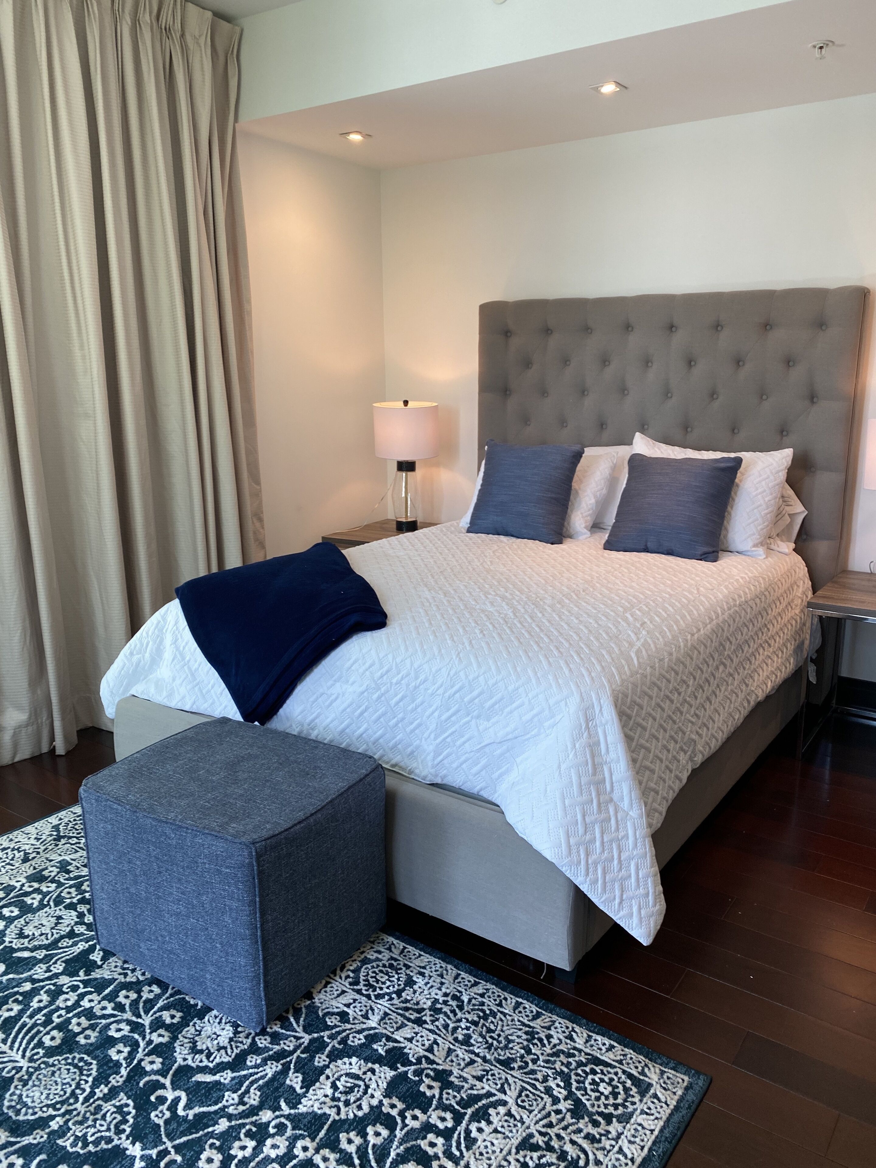 A neatly arranged bedroom with a large bed, a bedside table with a lamp, a grey ottoman, and a patterned rug.