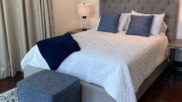 A neatly arranged bedroom with a large bed, a bedside table with a lamp, a grey ottoman, and a patterned rug.