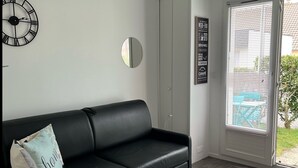 A modern living room with a black leather sofa, a round mirror, a wall clock, and a door leading to a balcony.