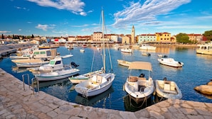 A marina with various boats docked, a stone quay, and buildings in the background.