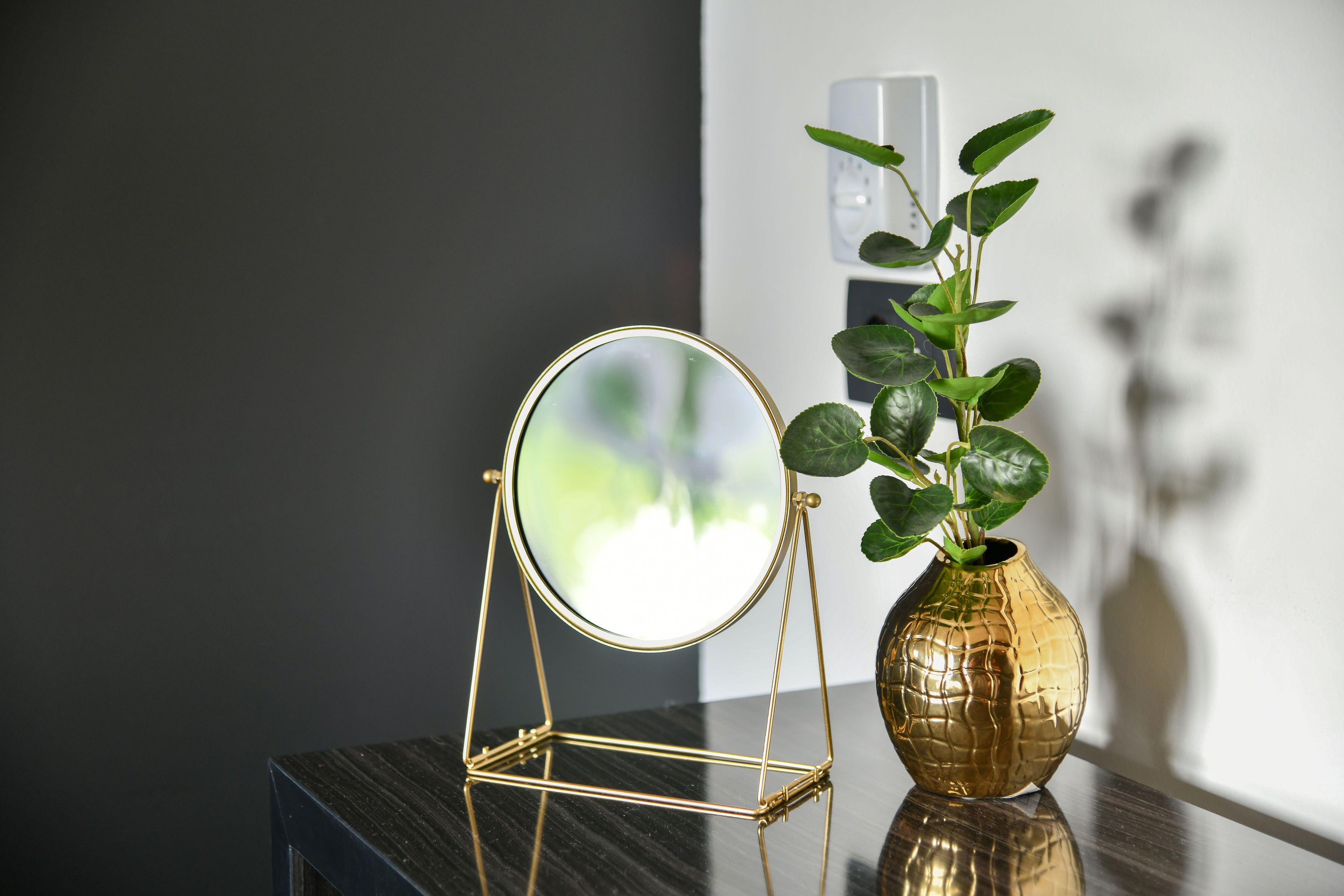 A round mirror on a stand, a gold vase with a plant, and a marble table against a dark wall.