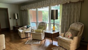 Een woonkamer met houten vloer, twee fauteuils, een bank, een bijzettafeltje met een lamp, en uitzicht op een bakstenen gebouw door grote ramen.