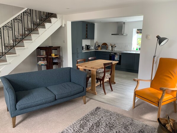 Ein Wohnzimmer mit blauem Sofa, einem Holzespeisetisch mit Stühlen, einer Treppe mit schwarzen Geländern und einer Küche im Hintergrund.