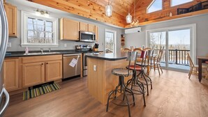 A spacious kitchen with wooden cabinets, a central island with bar stools, and large windows offering views of the outdoors. - Deer Crossing Retreat - new home close to Saddleback (Dallas Plantation)